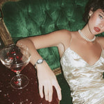 Woman in a white dress sitting on a green velvet couch with a glass of red wine.
