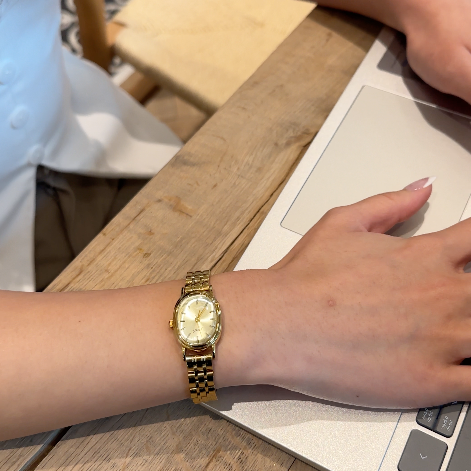Person wearing a gold watch on a wooden table with a laptop