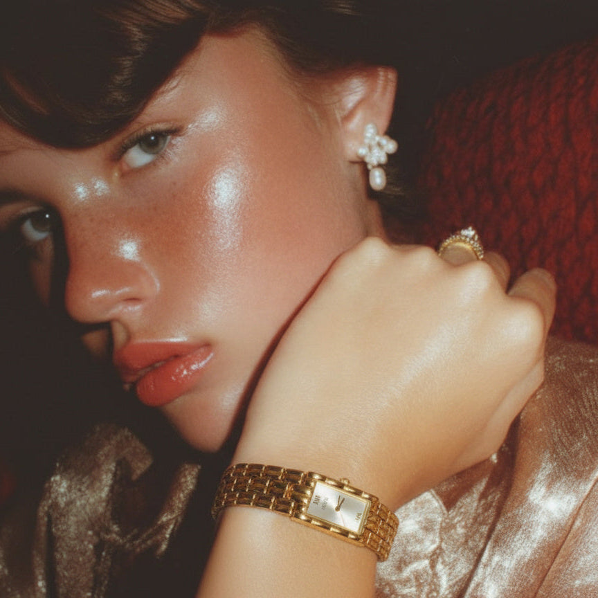 Woman wearing gold jewelry including a watch and ring, with a red background.