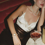 Woman in a white top and black sequin skirt sitting at a table with a drink, wearing jewelry.