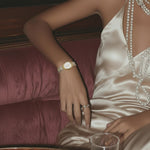 Woman in a satin dress with jewelry sitting on a couch next to a glass of whiskey.